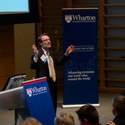 A person in business attire is speaking at a podium with Wharton branding in the background, likely at an educational or professional event.