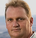 Headshot of a person with short, light hair, looking directly at the camera. The background is blurred, suggesting an outdoor setting.